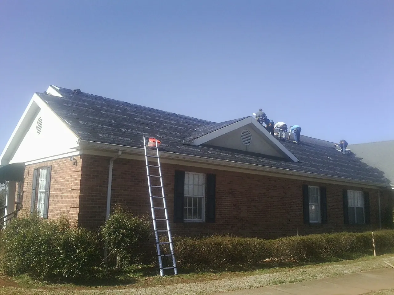 Crew tearing shingles off a brick ranch roof with a ladder set — Metal Roof Repair work by our crew in Bellville