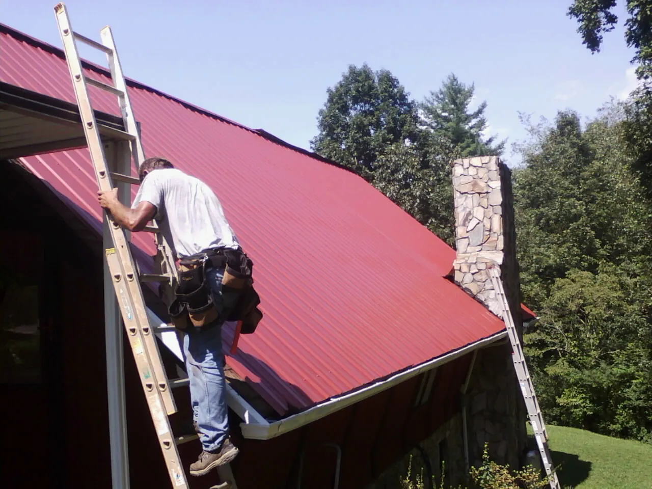 Roofer on a ladder beside a red metal roof and stone chimney — Soffit & Fascia Repair by Roofing Brothers in Bellville